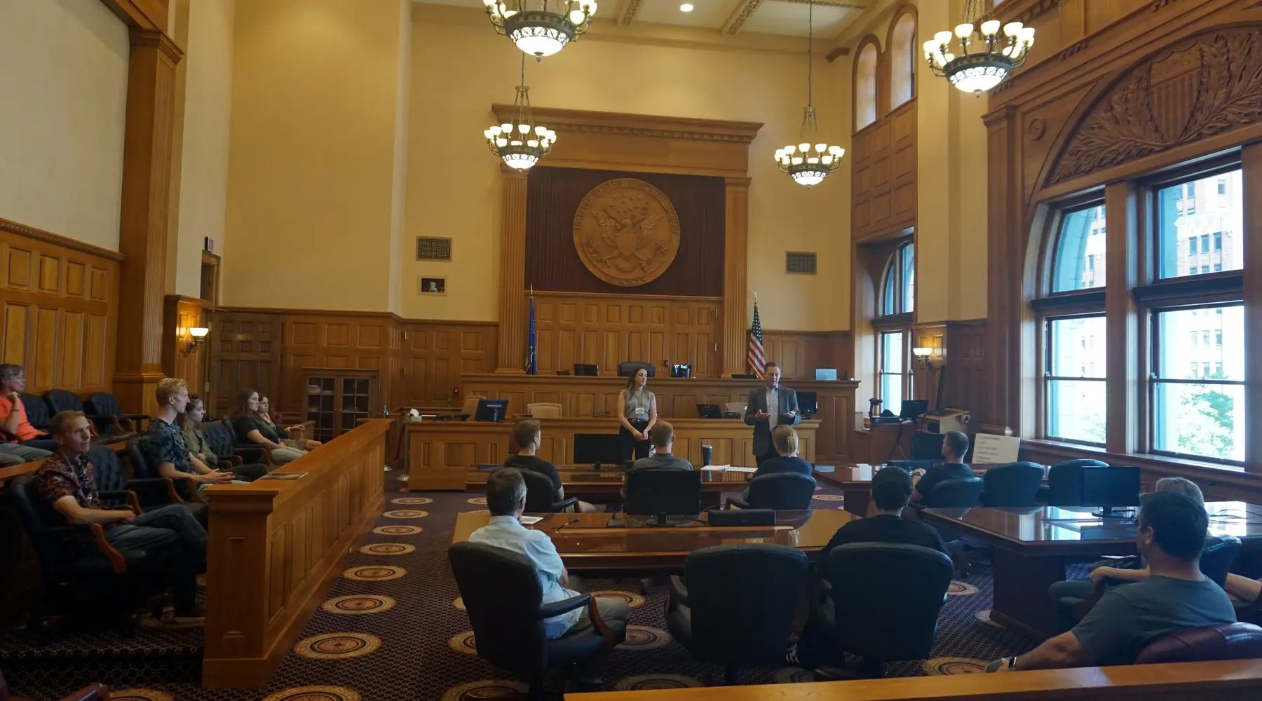 An attorney speaking at the lectern of a United States federal courtroom while the judge sits on the bench and the audience watches.